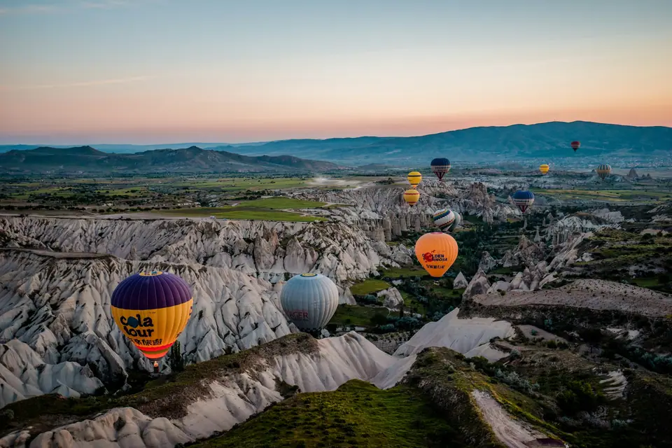 Dozens of hot air balloons rising at sunrise over the rock cones and ridges of a Cappadocia valley, seen from inside a wicker basket