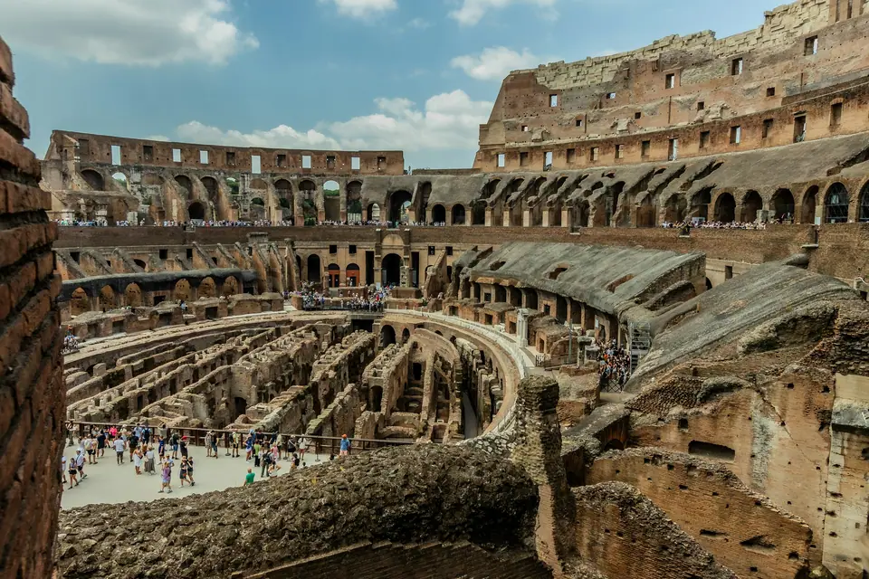 Brick corridors and stone walls of the Colosseum hypogeum seen from above, visitors on a walkway between them, the tiers of arches rising behind