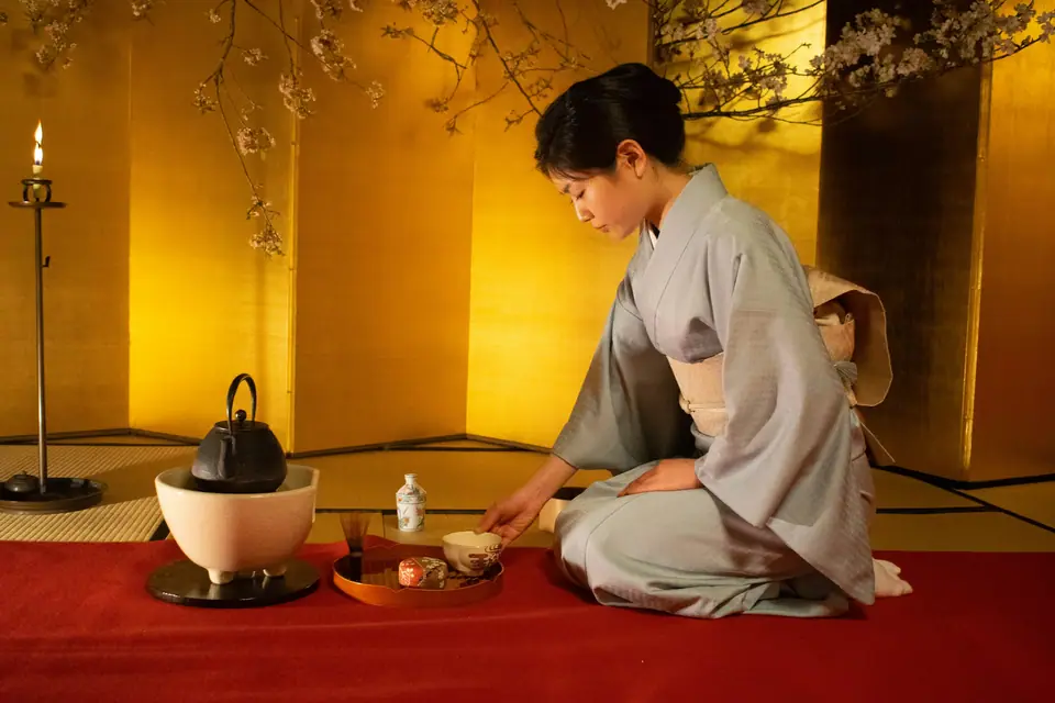 Host in kimono whisking matcha in a black ceramic bowl on a tatami floor, a hanging scroll and a single flower in the alcove behind