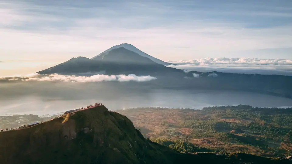 Hikers in jackets sitting on the dark volcanic rim of Mount Batur at dawn, Lake Batur below in the caldera and the cone of Mount Agung above a sea of cloud