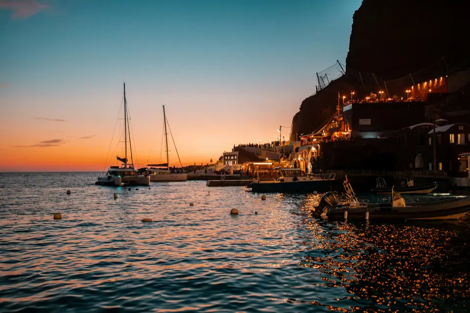 White sailing catamaran anchored in the Santorini caldera at sunset, passengers on the front nets, dark cliffs and white villages above