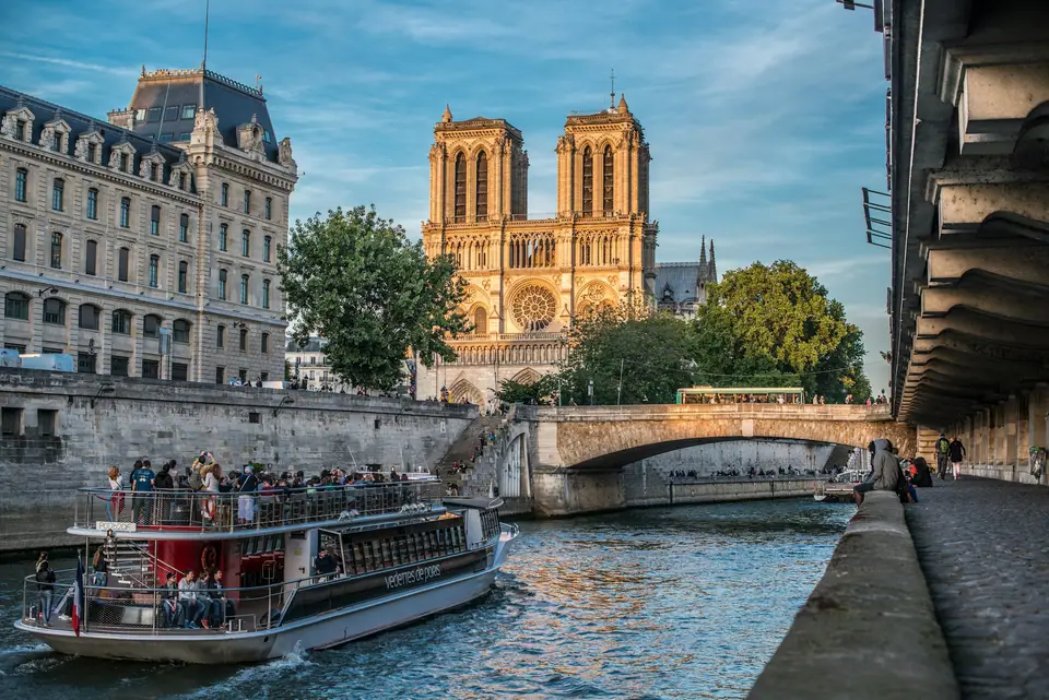 Open-top sightseeing boat on the Seine at dusk, passengers on the deck, the Eiffel Tower lit up behind a stone bridge