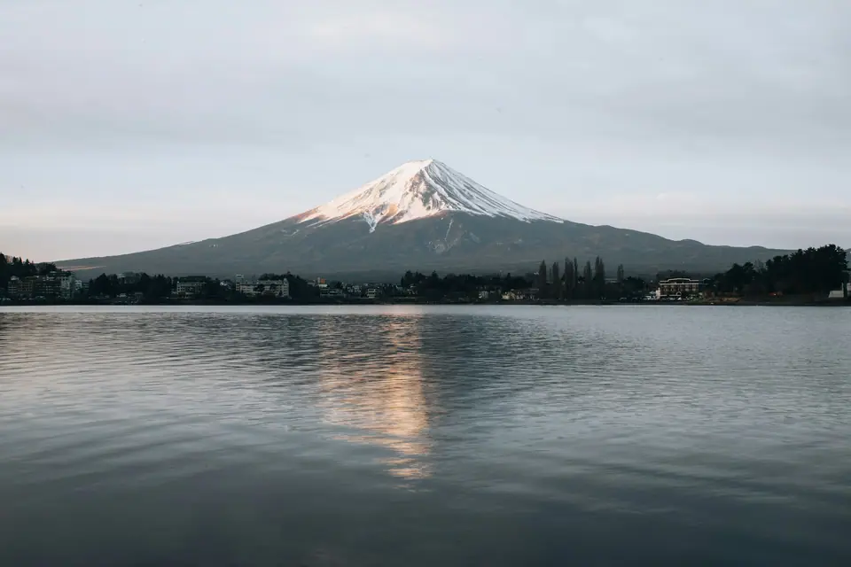 Mount Fuji with a cap of snow rising above Lake Kawaguchi, its reflection on the calm water and autumn trees along the shore