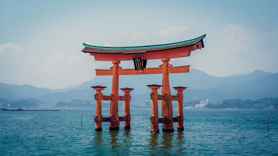 The large vermilion torii gate of Itsukushima Shrine standing in the sea off Miyajima at high tide, with forested hills in the background