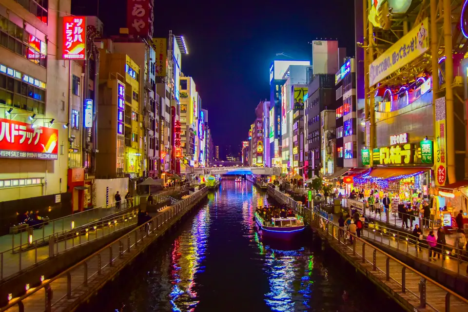 The Dotonbori canal in Osaka at night, lined with giant neon billboards including the Glico running man sign, with crowds on the Ebisubashi bridge