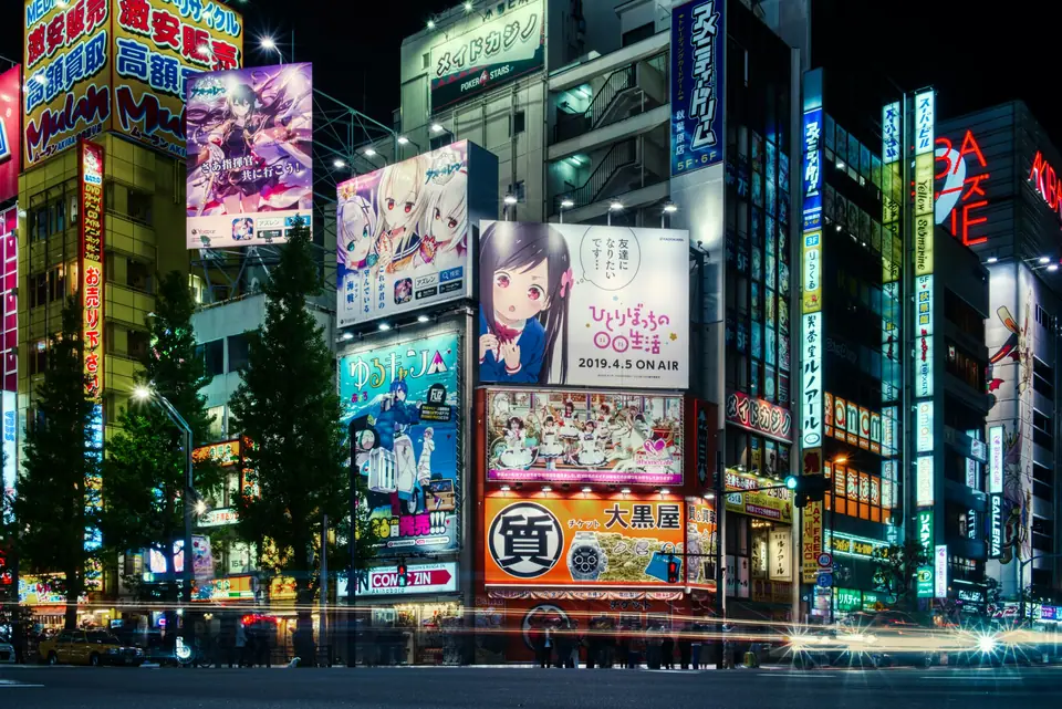 A Tokyo street at night in Shinjuku, lit by stacked neon and LED signs, with pedestrians crossing under the lights and a tall office tower behind