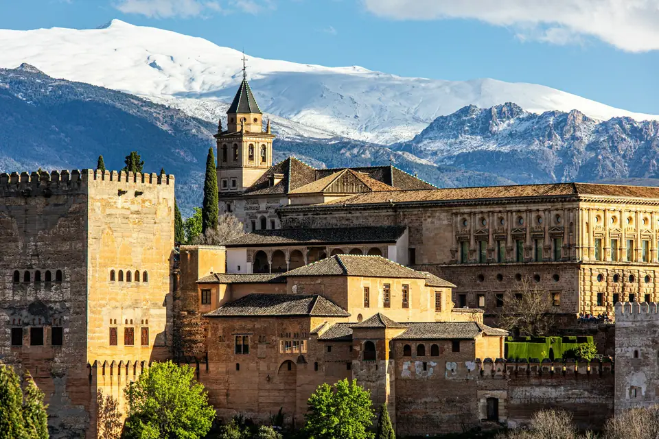 The Alhambra palace on its wooded hill above Granada, its red walls and towers lit by late sun, with the snowy peaks of the Sierra Nevada behind