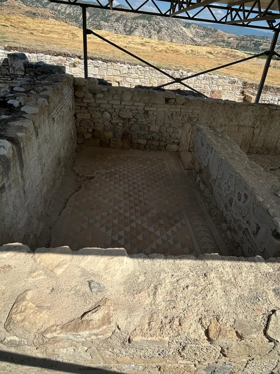 Room with a geometric mosaic floor under a modern steel shelter roof in the Mosaic House at Tripolis